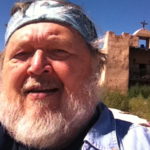 marc severson stands in front of an old style spanish mission. he is bearded and wear a bandana on his head