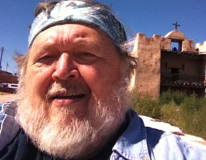 marc Severson stands in front of an old style Spanish mission. he is bearded and wear a bandana on his head