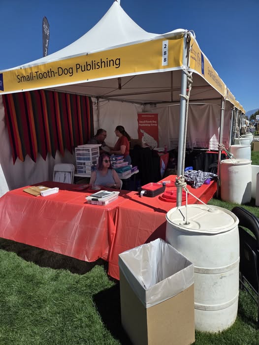 our booth at TFOB. white tent with yellow sign with name of company. red tablecloths and art supplies and booth edges.